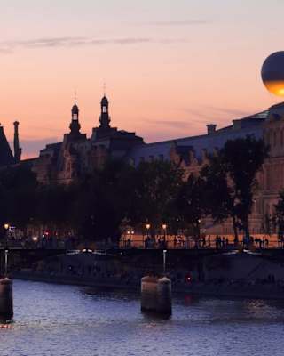 A sunset view of a river in Paris with silhouetted historic buildings, a bridge, and a large art installation resembling a white sphere on a rooftop, softly lit against the evening sky.