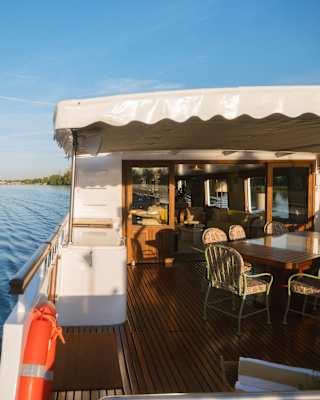 The covered deck of a boat with a dining table and chairs, moving along a calm river.