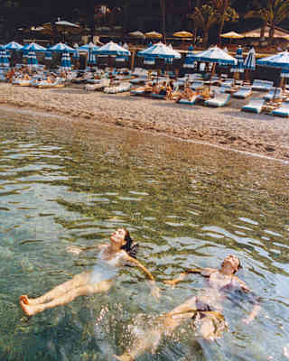 In front of the beachfront parasols, two women in swimming costumes sit in the shallows, heads tipped back, seen from above.