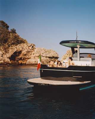 A couple sunbathes on the lounge deck of an Apreamare Gozzo boat as sails in full sun towards the headland, seen from behind.