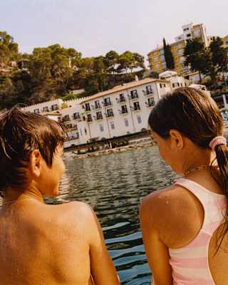 Angled image of a young girl and boy, wet from swimming, sitting in sun on a boat facing Mazzaro beach, viewed from behind.