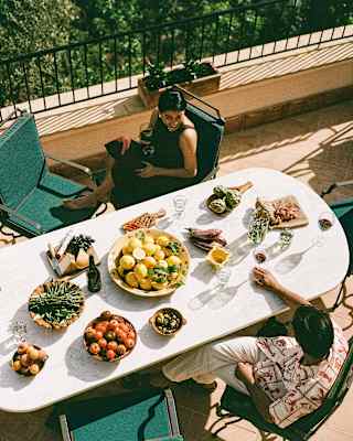 A couple sit opposite each other on green chairs at a lunch table with salads, cheeses, breads and wine, seen from above.