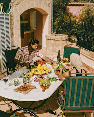 A couple sit in half-sun at a large outdoor table covered in dishes of charcuterie, bread and fresh produce, with red wine.