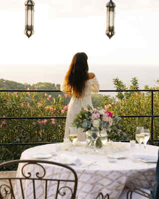 A dark-haired woman in white dress gazes at the view from terrace, seen over a table where a male companion sits with wine.