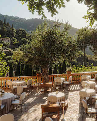 View across a stone terrace with a lush hillside aspect, where white-cloth dining tables await guests in dappled sunshine.