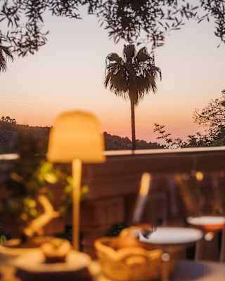 A cosy outdoor table with wine glasses and a lamp overlooks a sunset view with palm trees and distant hills.