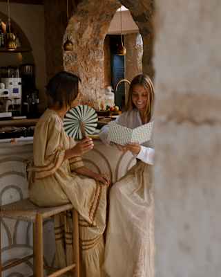 A female guest fans herself as another looks at a menu, seated on stools at Tramuntana Grill, seen through the stone arches.