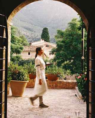 A white-shirted waiter in a long apron tied at her waist walks along the garden terrace, framed by an ancient stone arch