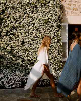 A female guest with long blond hair, wearing a white linen wrap dress strides past the jasmine-clad hotel, seen side on.
