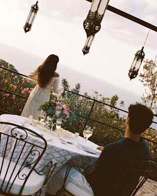 Angled image of a man at a dining table as he gazes at a woman in a white dress with long dark hair, standing on the terrace.
