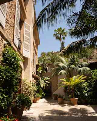 Palm frond shadows play on the stone-tiled, plant-filled terrace outside a residence with white shutters and stone walls.