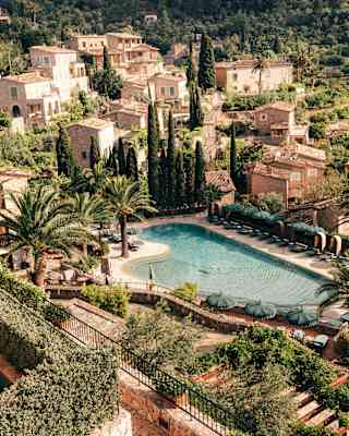 Aerial view of the tropical-shape pool bordered by dark-green parasols and palm trees, overlooked by rustic stone residences.