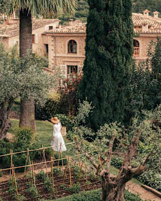 A woman in a white dress with a hat and straw basket wanders along the Poet's Walk path through gardens, seen from above.