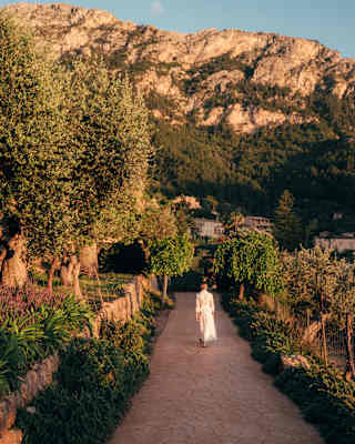 A woman in a white dress walks along a path between gardens held back by stone walls and borders, seen in golden light.