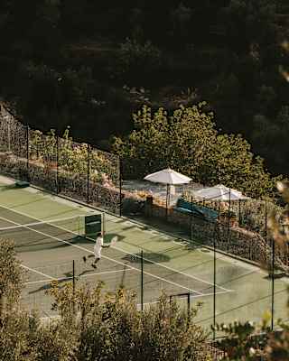 Two men play tennis on a green court surrounded by trees and fencing, with two white courtside parasols, seen from above.