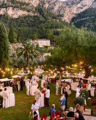 The Tramuntana Mountains rise behind the hotel gardens where guests gather beneath pretty light strings at an evening event.