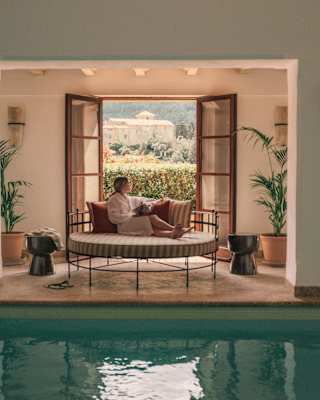 A woman in a robe reclines on a day bed gazing back at the garden view through open doors behind her, seen over the spa pool.