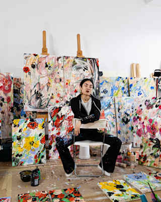Artist Anna Deller-Yee, in black, sits astride a chair, arms on the backrest in a studio filled with vibrant floral artworks.