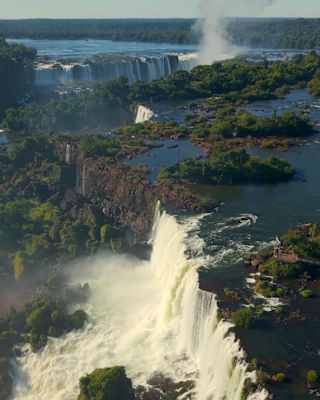 Aerial view of the Iguassu falls with creamy rapids cascading over the forested rocky edge, with more falls in the background.