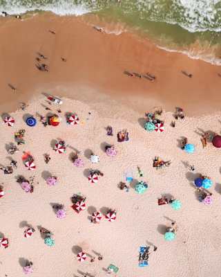 Aerial shot of a pinkish sand beach dotted with sunbathers and candy-like parasols in mint, pink, and red-and-white stripes.