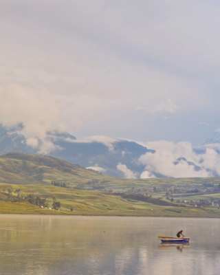 Snowy peaks rise on the horizon in a view over Laguna Huaypo, where a man tends to a boat on the still, green-tinted waters.