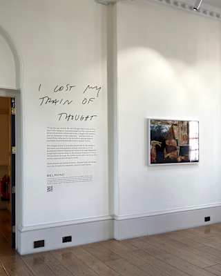 A white wall displays text reading “I lost my train of thought” above a block of print in an airy gallery with wood floors.