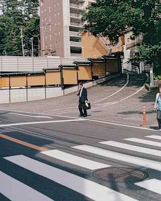 A businessman in a dark suit  with a briefcase walks from blocks of apartments towards a wide road with crossing markings.