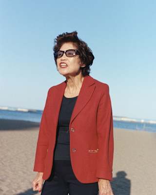 A Japanese woman with short black hair, wearing dark glasses and clothes with a red jacket, stands on a wide sunny beach.