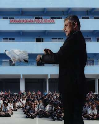 A magician in a black suit releases a white dove in front of a seated children in front of Gnanabhaskara Public School.