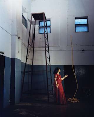 A woman with dark hair and a red dress levitates a rope so it stands high above her head by a tall platform in a large room.