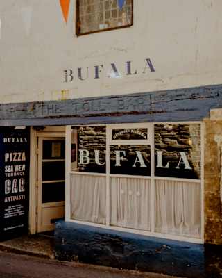 The exterior of a building with a sign reading BUFALA. There are string bunting above, a window with curtains, and a blackboard advertising pizza near the entrance. The building façade appears weathered.