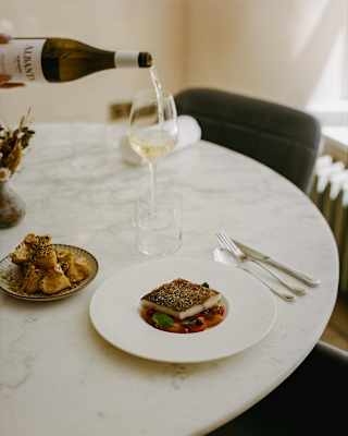 A hand pours white wine into a glass on a round marble table set with a plated fish dish, a small plate of bread, and cutlery.