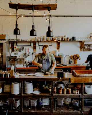 A chef stands behind a rustic kitchen counter, preparing food. Hanging lights and shelves with crockery and kitchenware are visible in a cosy, warmly lit restaurant setting.