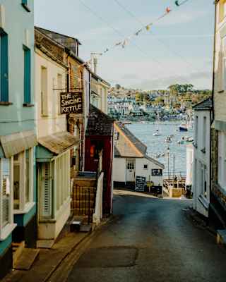 A narrow street leads downhill between colourful buildings toward a harbour filled with boats. String lights or flags hang above, and the sky is clear with soft sunlight.