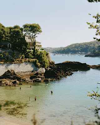 A small, rocky beach with a few people swimming in clear water. Trees and a house are on the left, overlooking the bay. Boats float in the distant water under a clear sky.
