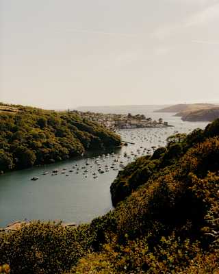 A scenic view of a river or bay with boats moored on the water, surrounded by green hills. A small town is visible in the distance under a clear sky.