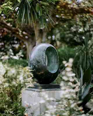 A smooth, oval-shaped abstract sculpture with a hollow centre stands on a stone plinth, surrounded by lush greenery and flowering plants in a garden.