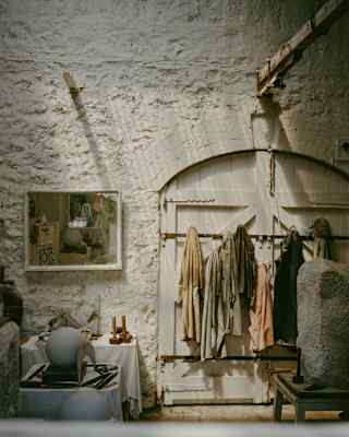 Clothing in neutral tones hangs on a rail against a textured white wall; a bench with cushions and a small table with decor items sit nearby, with soft natural light illuminating the scene.