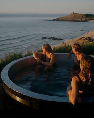 Three people relax in a round hot tub overlooking the sea and beach at sunset, with gentle waves and distant cliffs visible under a clear sky.