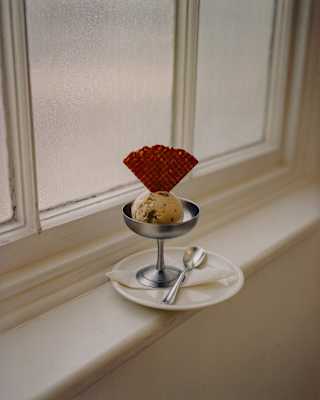 A metal cup with a scoop of ice cream and a waffle sits on a white plate by a window. A small spoon and fork rest on the plate.