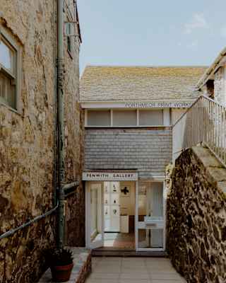 A narrow stone alleyway leads to the entrance of a small art gallery named Fountain Gallery. The building has stone walls, a shingled upper façade, and a potted plant by the door under a clear sky.