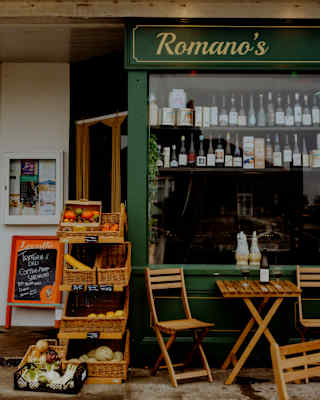 Small outdoor café with wicker baskets of produce, a chalkboard menu, wooden tables and chairs, and a green shopfront sign reading Romanos. Bottles and jars are visible in the window display.