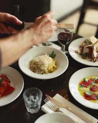 A person grates cheese over pasta at a table set with various dishes, a glass of wine, and cutlery. The scene suggests a freshly prepared meal in a cosy dining setting.