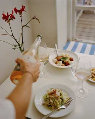A hand pours rosé wine into a glass on a white table set with plates of pasta, salad, wine glasses, and a vase with red flowers.