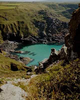 A turquoise cove surrounded by steep, grassy cliffs under a clear sky, with rocky terrain leading down to the water.