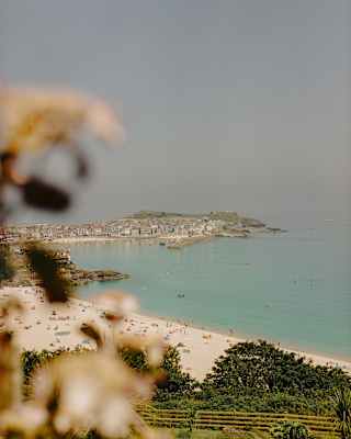 A coastal town with sandy beaches, turquoise water, and distant hills under a hazy sky. Out-of-focus plants frame the foreground.