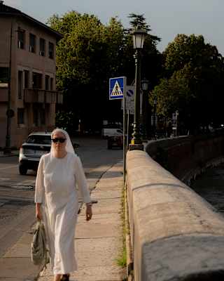 A woman in a white habit and sunglasses walks along a pavement beside a stone wall.