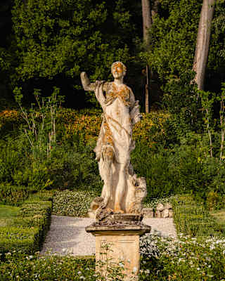 A weathered stone statue of a classical figure stands on a plinth in a lush garden.
