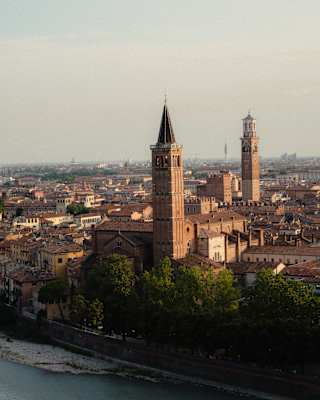 Aerial view of an old city skyline at sunset, featuring two prominent bell towers rising above terracotta rooftops and a river in the foreground.