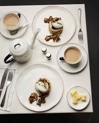 View down on two breakfasts of eggs on crumpets, with a dish of butter, a teapot and cups of tea, all served in white crockery.